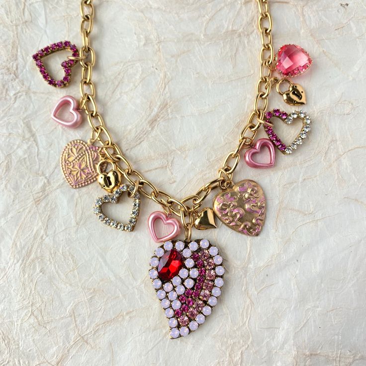 "The sweetest Valentine's Day charm necklace filled with an alluring mix of dangling heart charms. Heart charms include classic brass heart charms, some with shimmering Swarovski rhinestones, playful little pink hearts, and a bold rhinestone collage heart focal point to steal the show. These heart charms wire wrapped to perfection dangle from a high-quality gold-plated cable chain. This necklace has a solid and nice heavy weight to it for durability. 18\"l with 3\" extension. Lobster claw closure. Handmade in USA. ~NOTE: Some of the small heart charms are upcycled vintage pieces with limited quantity in our supply so they may vary from the photo but we are confident you'll love them!" Valentine's Day Vintage Charm Necklaces, Vintage Charm Necklace For Valentine's Day, Heart Shaped Vintage Charm Necklace For Valentine's Day, Valentine's Day Heart Pendant Necklace With Vintage Charm, Valentine's Day Metal Charm Necklace With Heart Pendant, Valentine's Day Heart Metal Charm Necklace, Valentine's Day Heart-shaped Metal Charm Necklace, Vintage Charm Heart Pendant Necklace For Valentine's Day, Vintage Charm Heart Pendant For Valentine's Day