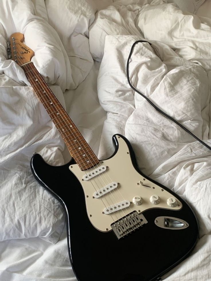 a black electric guitar laying on top of a white bed covered in sheets and pillows