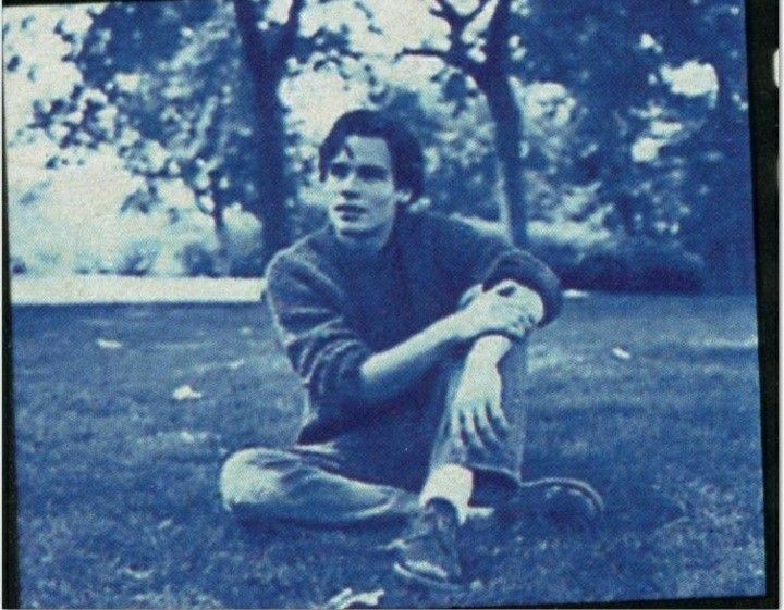 a young man sitting on the ground with his arms crossed and legs crossed, in front of trees