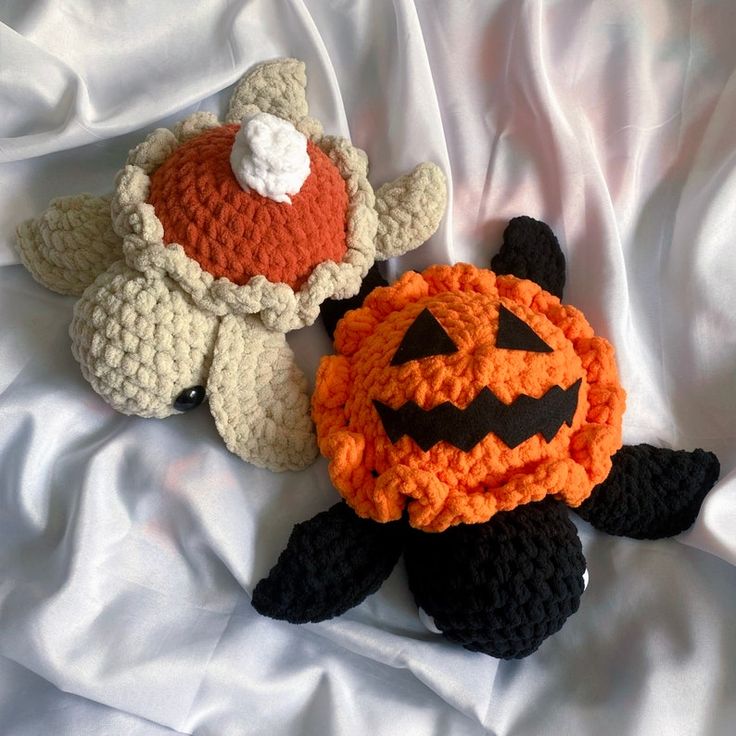 two crocheted stuffed animals laying on top of a white bed sheet, one is wearing a pumpkin hat and the other has a jack o lantern