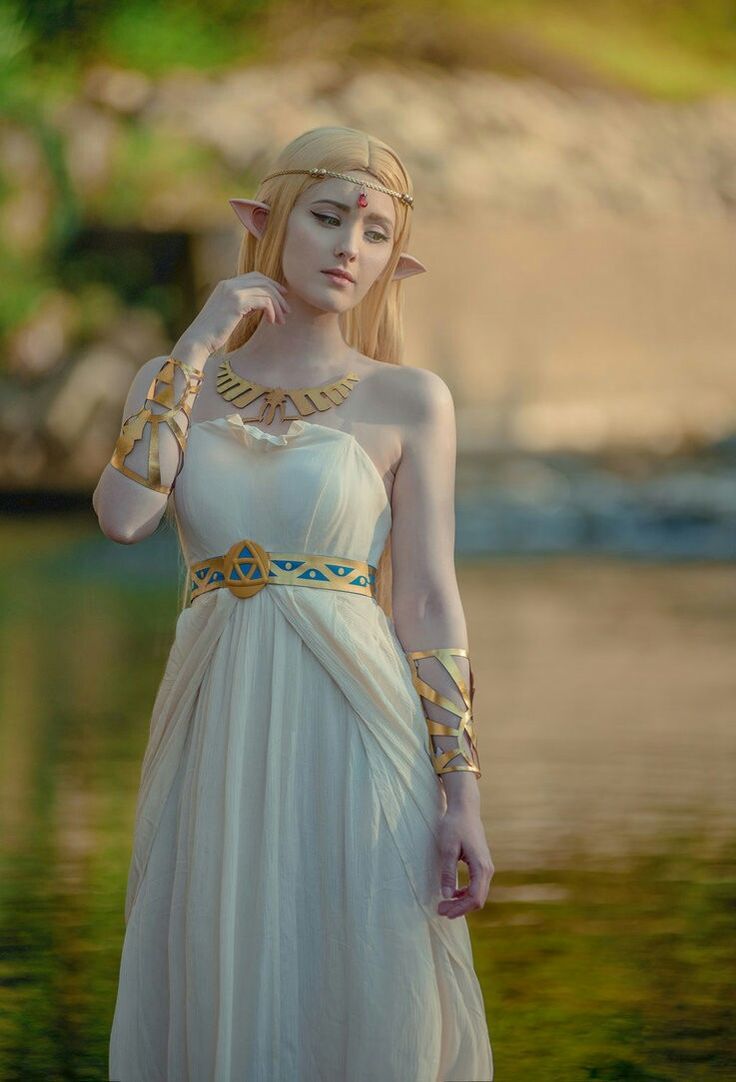 a woman in a white dress with gold jewelry on her neck and hands near her face