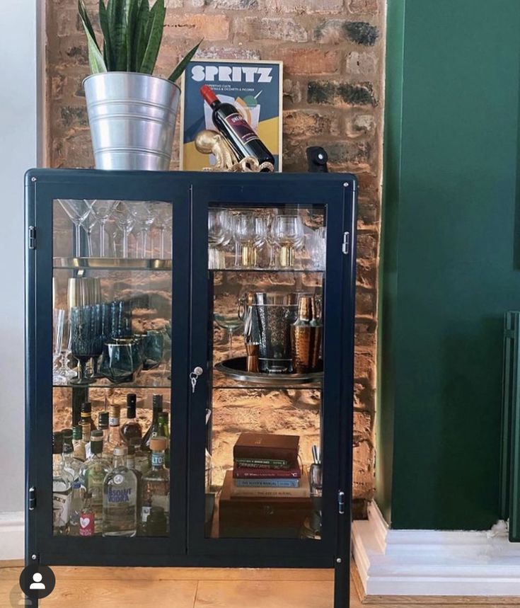 a glass cabinet with bottles and glasses on top in front of a brick wall next to a potted plant