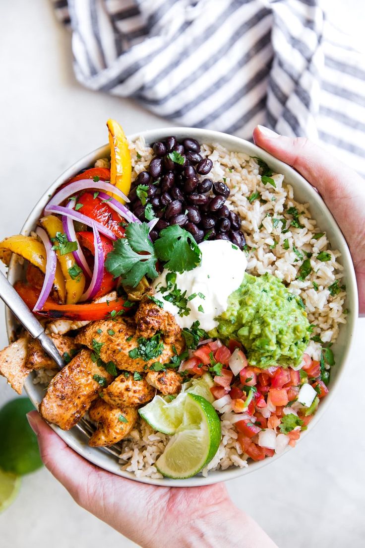 two hands holding a bowl of food with rice, beans, and avocado