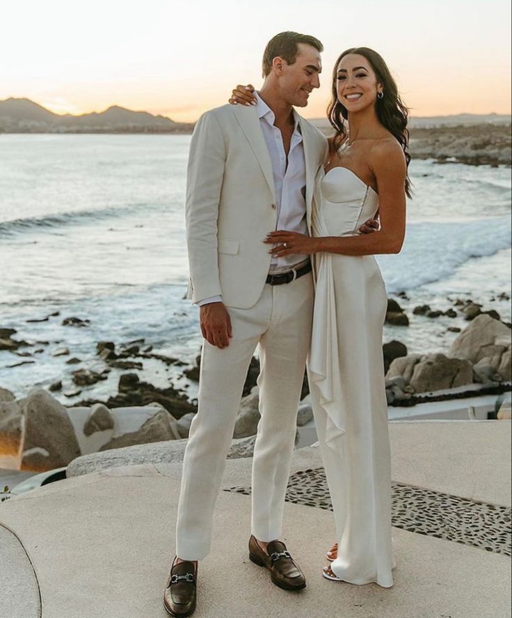 a man and woman standing next to each other in front of the ocean at sunset