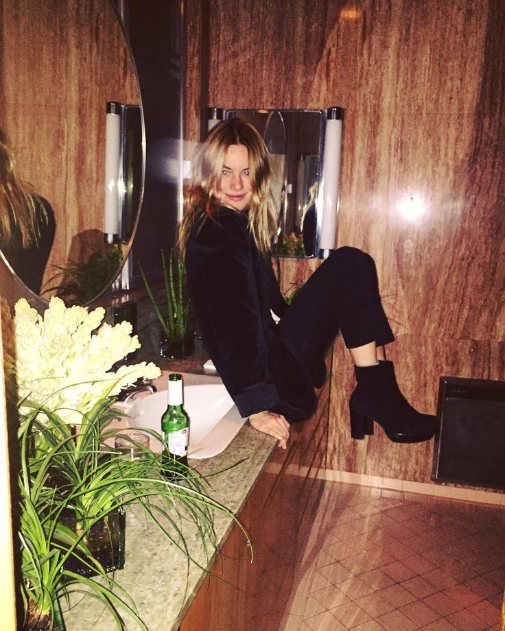 a woman sitting on a counter in a bathroom next to a plant and bottle of beer