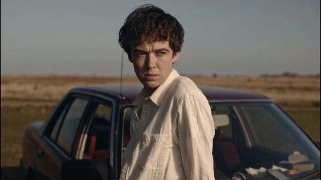 a young man standing next to a parked car in front of a field with grass