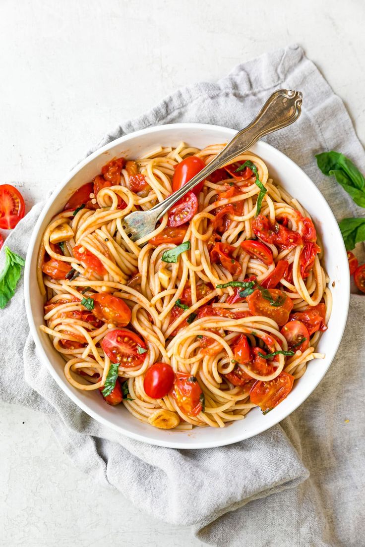 a white bowl filled with pasta and tomatoes on top of a cloth next to a fork