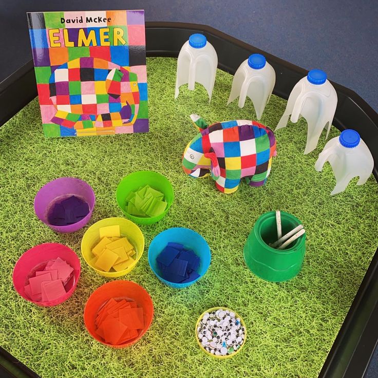 a tray filled with plastic cups and toys on top of green grass next to a book