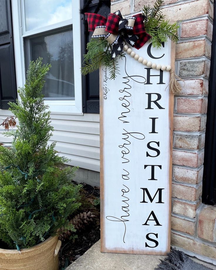 a wooden sign that says christmas is hanging on the side of a house with potted plants next to it