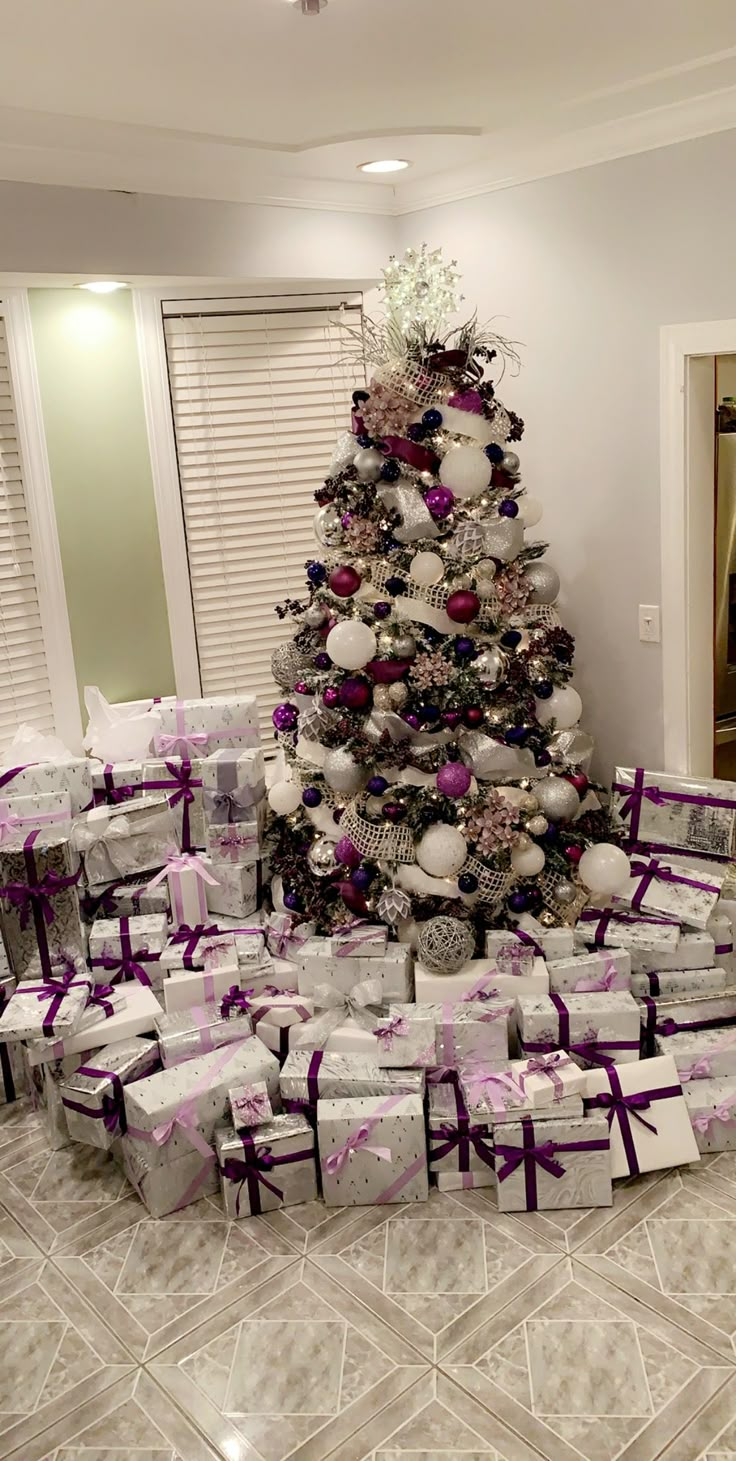 a christmas tree with presents under it in front of a wall decorated with purple and white ornaments