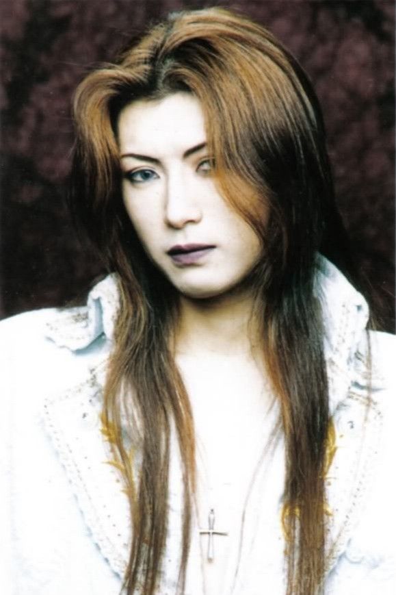 a woman with long hair wearing a white shirt and cross on her chest, in front of a brown background