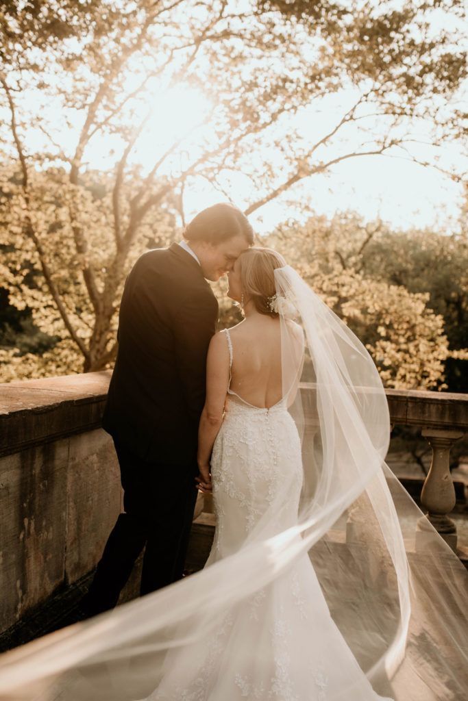 a bride and groom standing on a bridge with their veil blowing in the wind at sunset