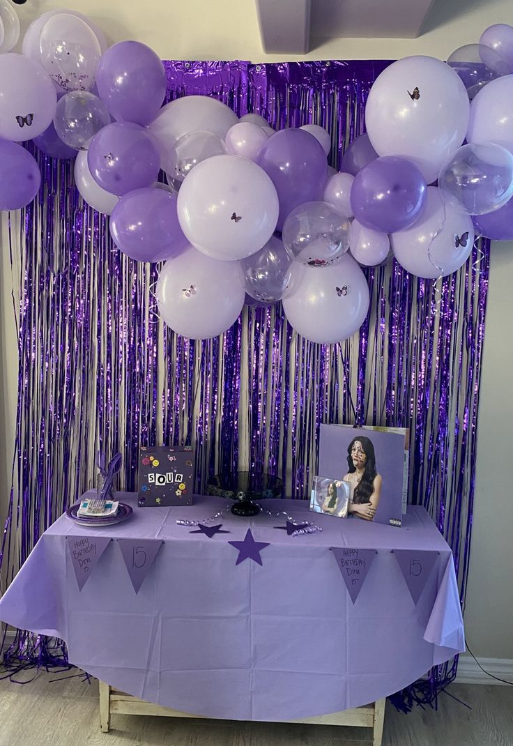 a table with purple and white balloons hanging from it's ceiling, next to a party backdrop