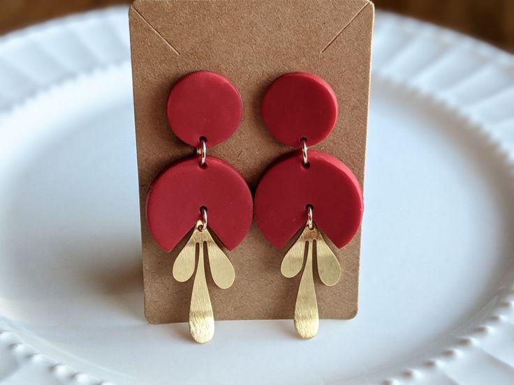 a pair of red and gold earrings sitting on top of a white plate next to a brown card