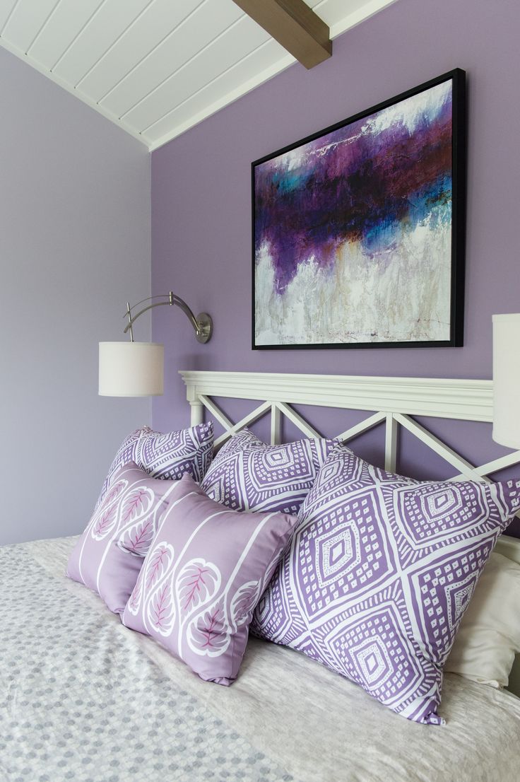 a bed with purple and white pillows in a bedroom next to a framed painting on the wall