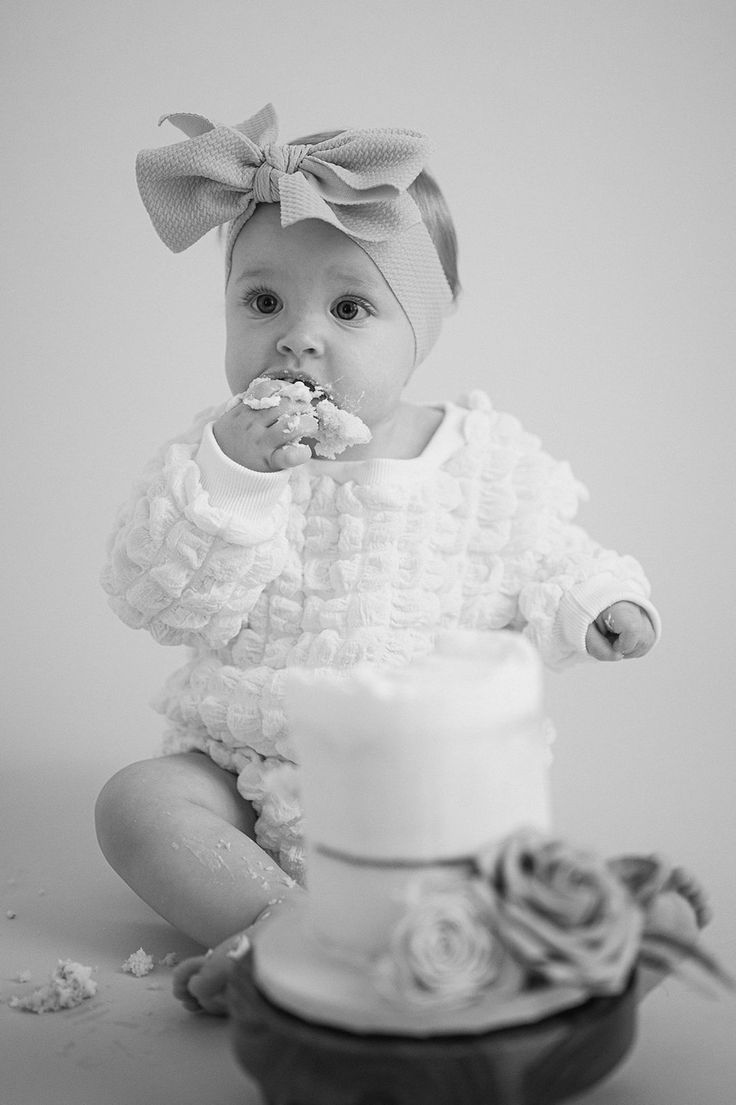 a black and white photo of a baby eating cake with a bow on her head