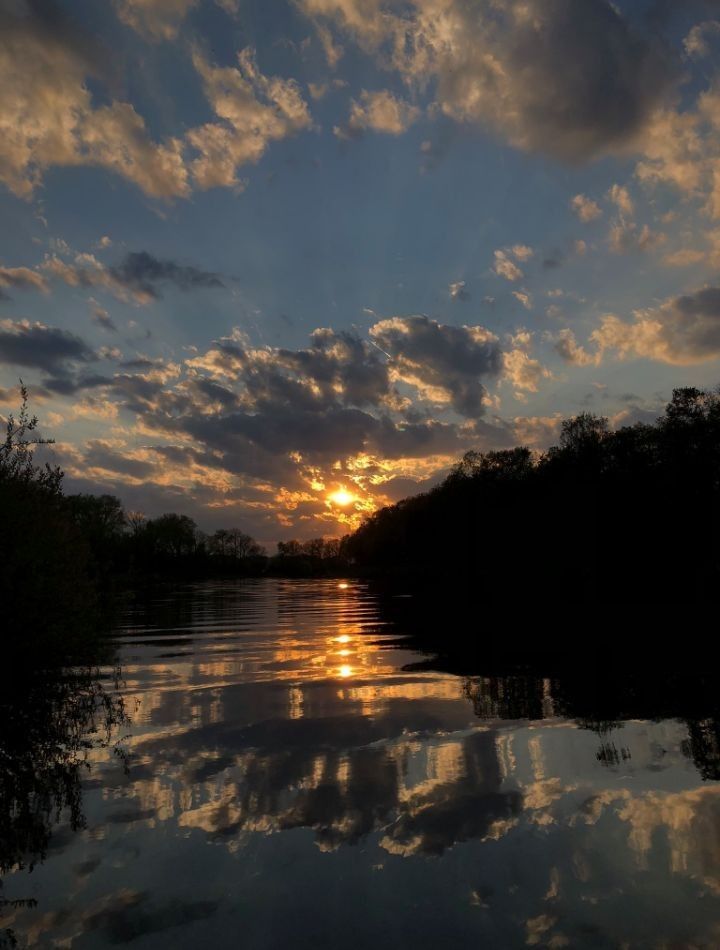 the sun is setting over water with trees in the foreground and clouds in the background