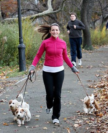 a woman walking two dogs on a leash down a path in the park while another man runs behind her