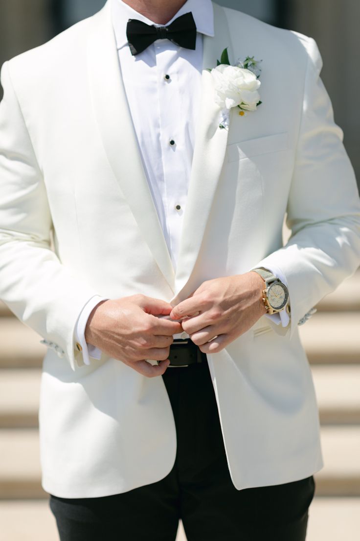 a man in a white tuxedo with a black bow tie and watch on his lapel