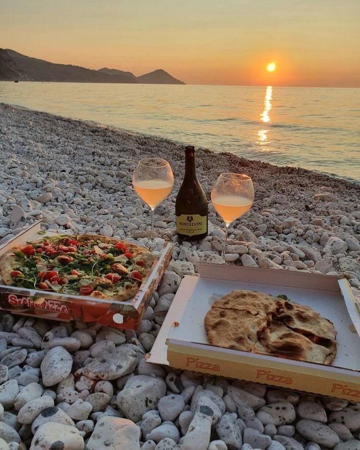 two glasses of wine are sitting on the beach next to pizza and water at sunset