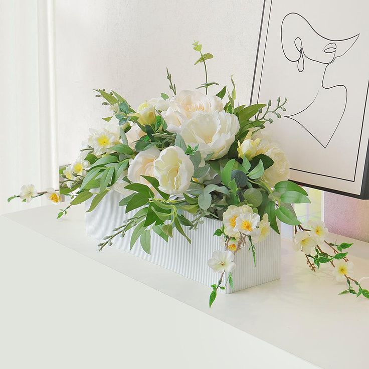 white flowers in a vase on a table next to a framed drawing and candle holder