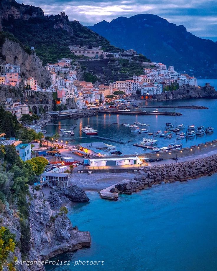 boats are docked in the water next to some buildings and cliffs at dusk, with mountains in the background