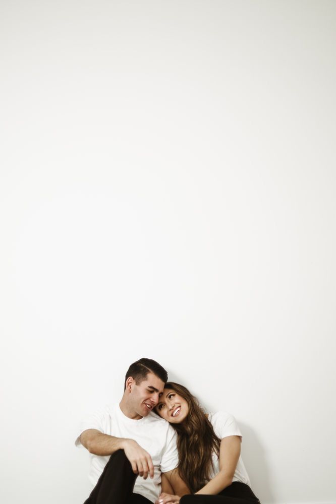 a man and woman sitting next to each other in front of a white wall with their arms around each other