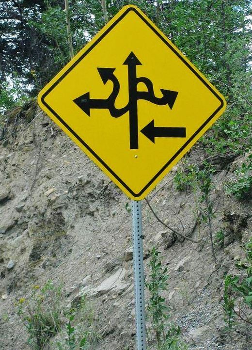 a yellow street sign with black arrows pointing in different directions on the side of a hill