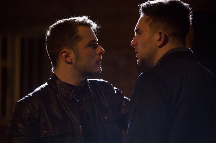 two men standing next to each other in a dark room with one looking at the other