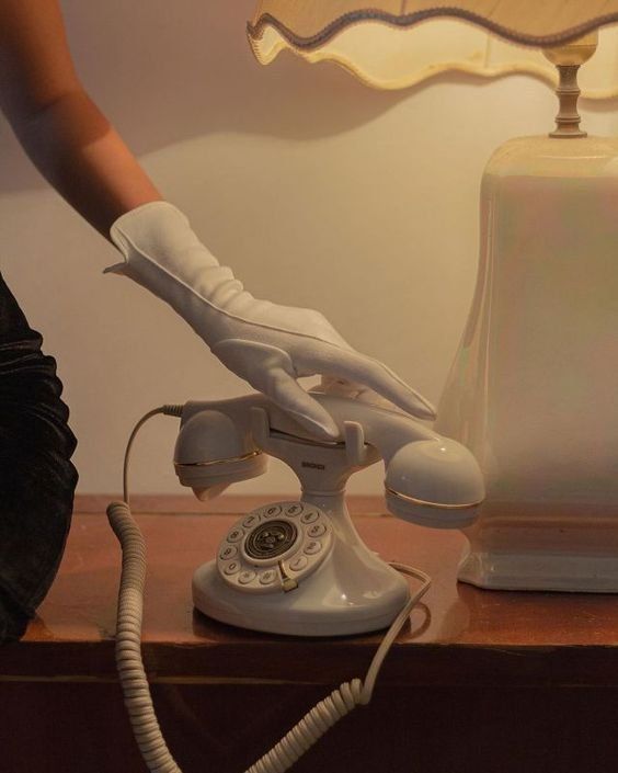 an old fashioned white telephone sitting on top of a wooden table next to a lamp