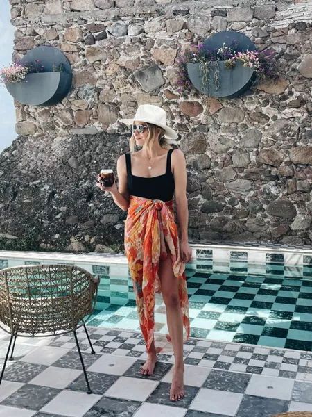 a woman standing in front of a swimming pool wearing a hat and dress with an orange sash around her waist