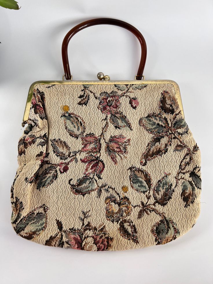 This bohemian, 1970s tapestry handbag is a true statement piece. The brown lucite top handle adds a touch of elegance, while the large fabric body is adorned with beautiful flowered branches and leaves on a light brown background. The tapestry design is sure to catch the eye and add a touch of vintage charm to any outfit. With a spacious 11 inches by 12 inches size, it's perfect for carrying all your daily essentials. Expected wear for a vintage item with minor surface wear on the frame. See pic Elegant Luxury Tapestry Bag, Vintage Tapestry Bag For Evening, Vintage Evening Bag In Tapestry Material, Vintage Cream Bags With Detachable Handle, Elegant Floral Print Top Handle Shoulder Bag, Vintage Tapestry Shoulder Bag For Evening, Vintage Tapestry Bag For Formal Occasions, Vintage Beige Satchel, Vintage Formal Tapestry Bag