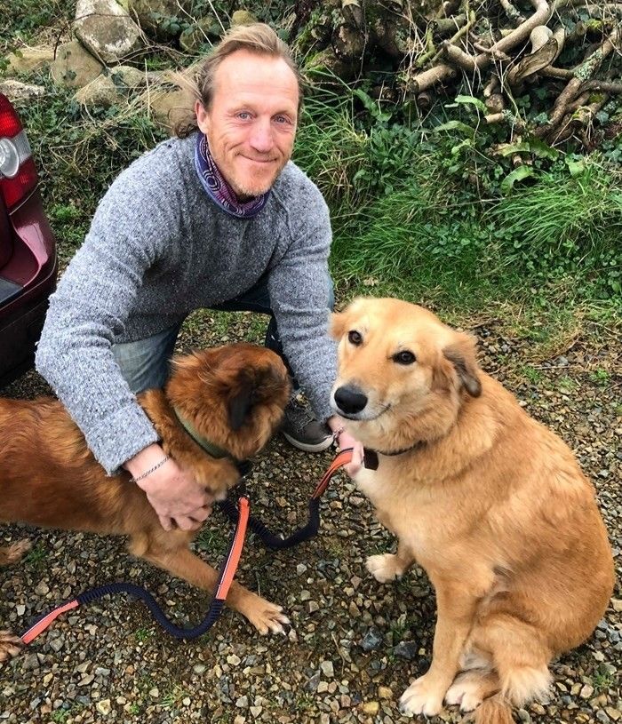 a man kneeling down with two dogs on leashes in front of him and a car behind him