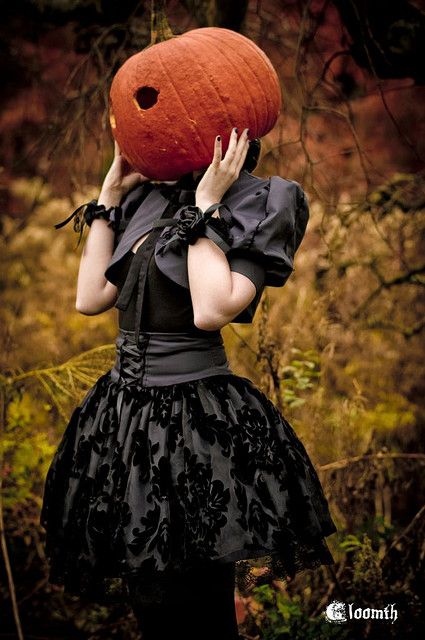 a woman in a black dress holding a pumpkin on her head and covering her face with both hands