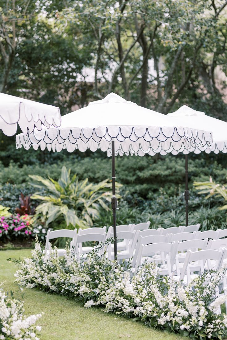 an outdoor ceremony setup with white chairs and umbrellas in the grass, surrounded by greenery