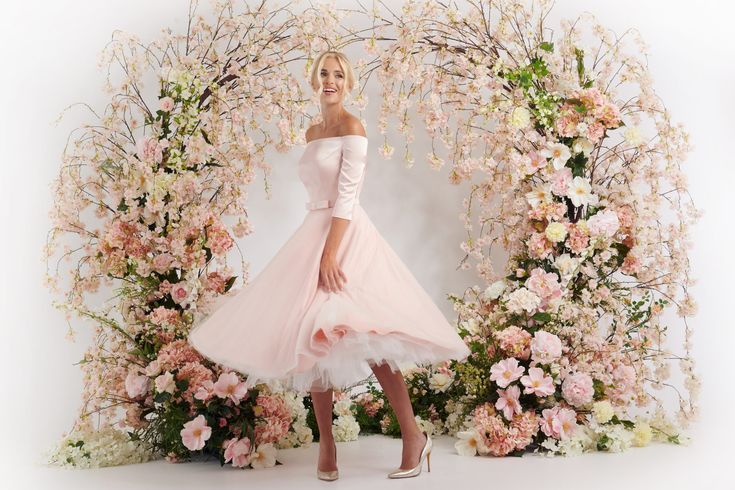 a woman in a pink dress standing next to a floral arch with flowers on it