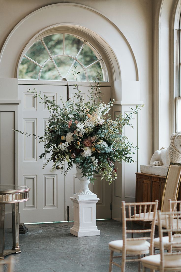 a vase filled with flowers sitting on top of a white pedestal next to chairs in front of a door