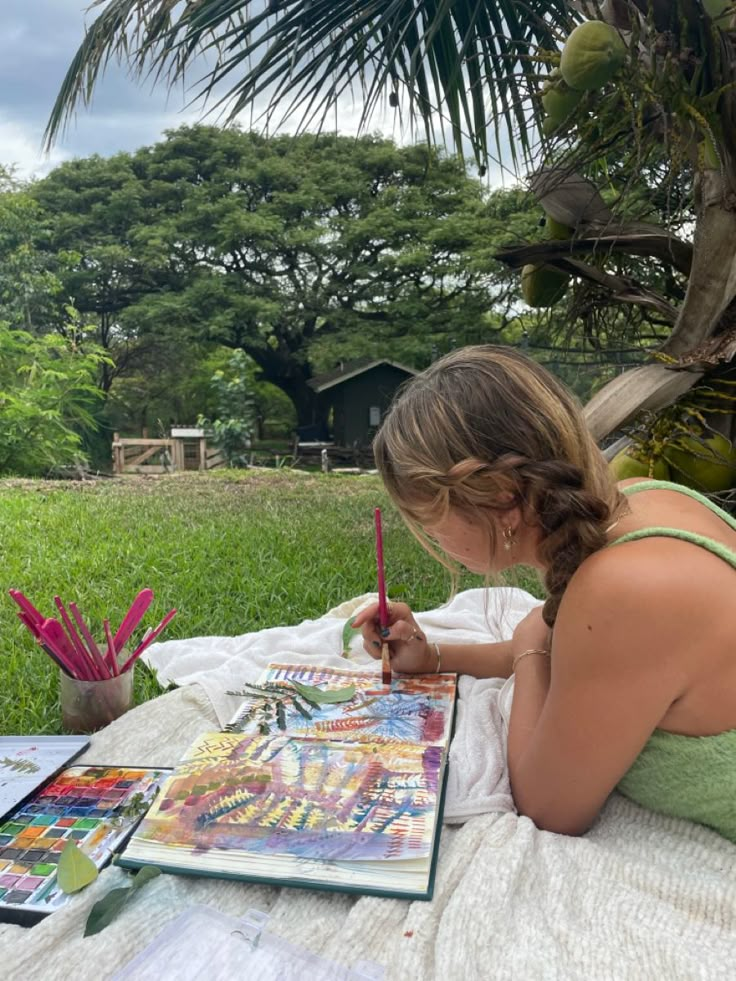 a woman sitting on top of a blanket next to a tree holding a paintbrush