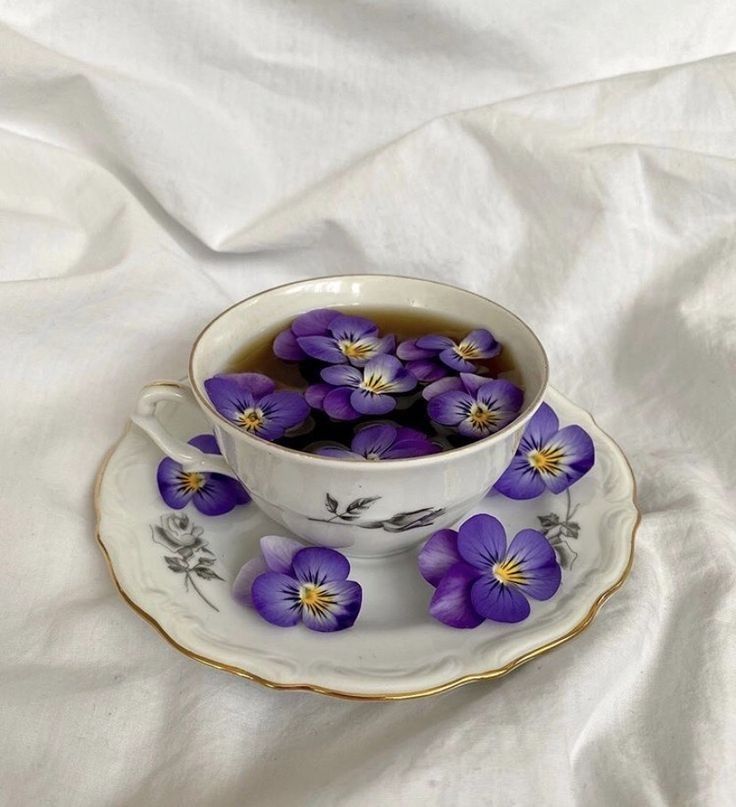 a tea cup filled with purple pansies sitting on top of a white table cloth