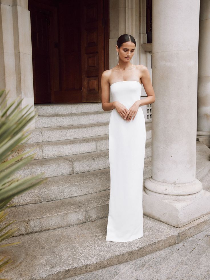 a woman in a white strapless dress standing on steps with her hands clasped to her chest