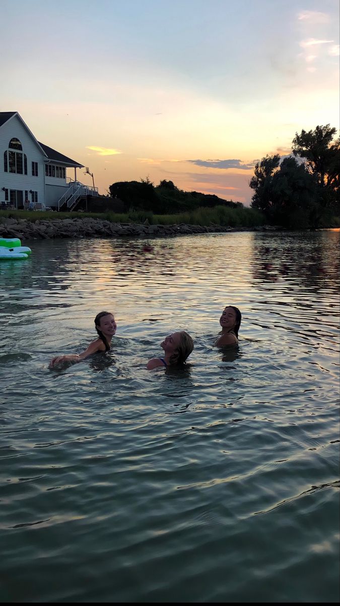 three people are swimming in the water near a house and boat at sunset or dawn