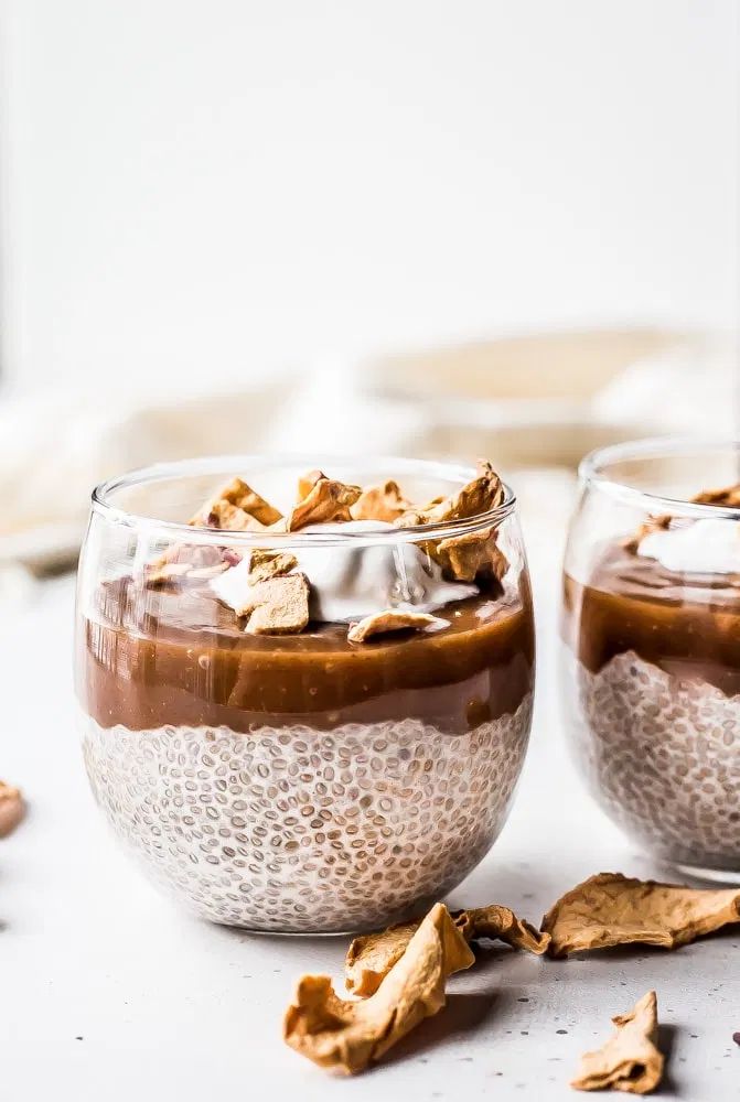 two glasses filled with dessert sitting on top of a table next to leaves and nuts