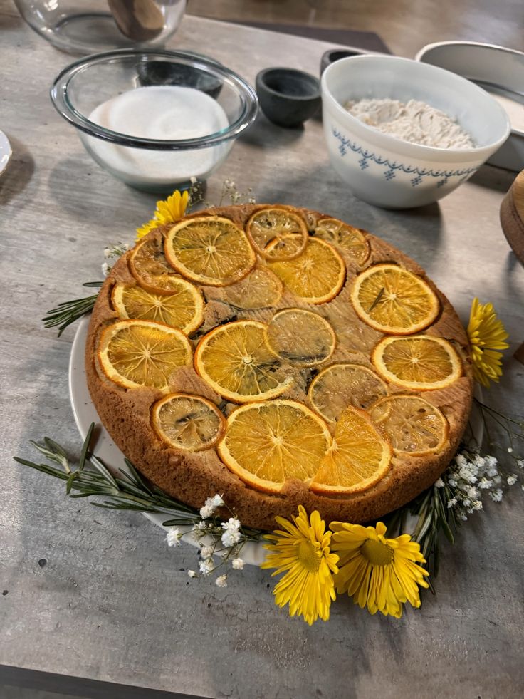 a cake with sliced oranges and flowers on a table next to other plates, bowls and utensils