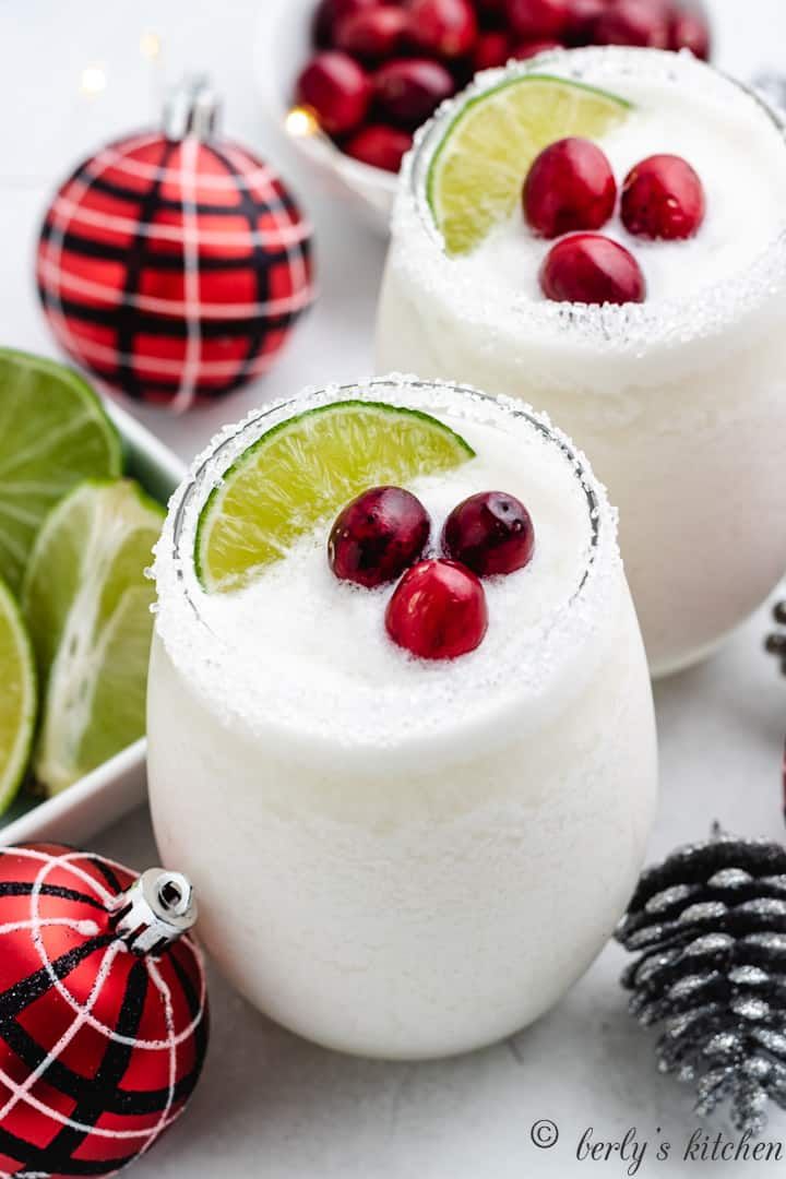 two margaritas with cranberries and lime on the rim next to christmas decorations