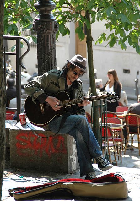 a man with a hat and sunglasses playing an acoustic guitar on the sidewalk in front of a cafe