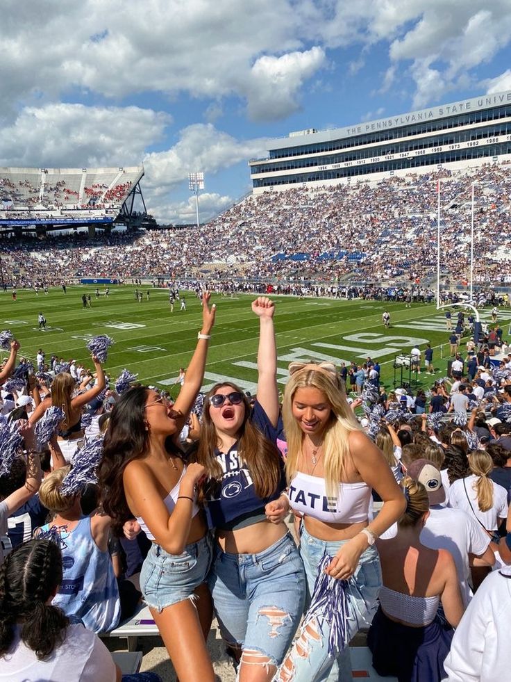 two women sitting in the stands at a football game with their arms up and one woman holding her hand up