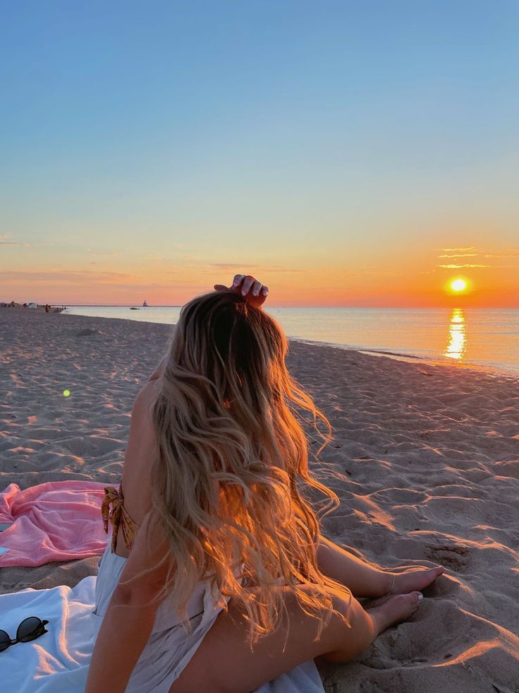 a woman sitting on top of a sandy beach next to the ocean at sun set