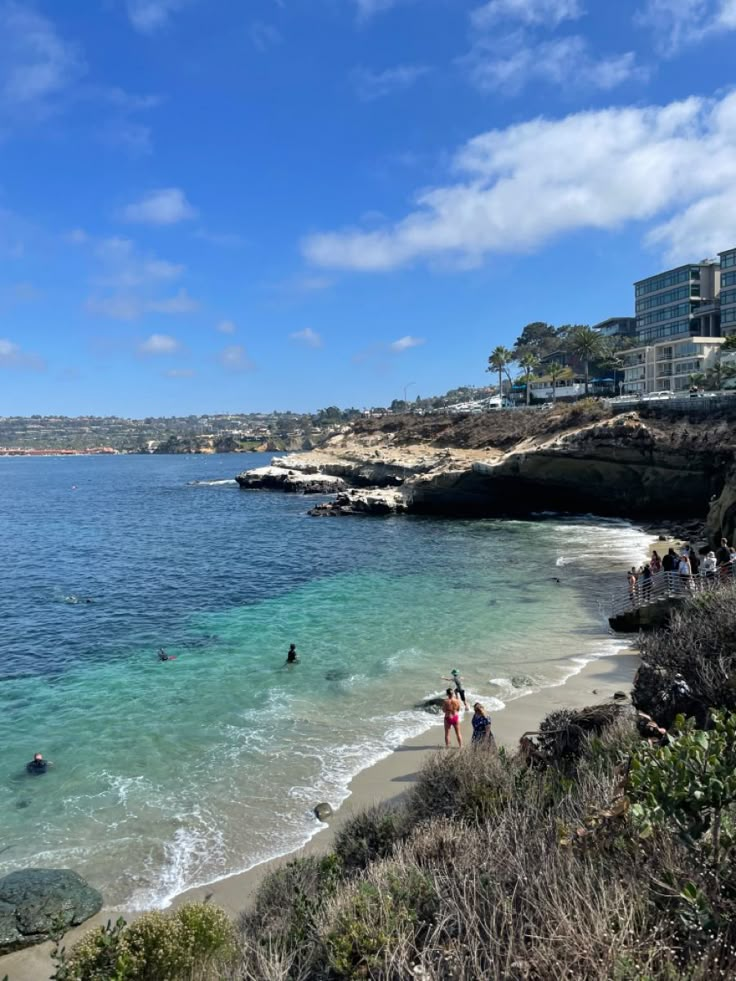 people are swimming in the water at a beach near some buildings and cliffs on a sunny day