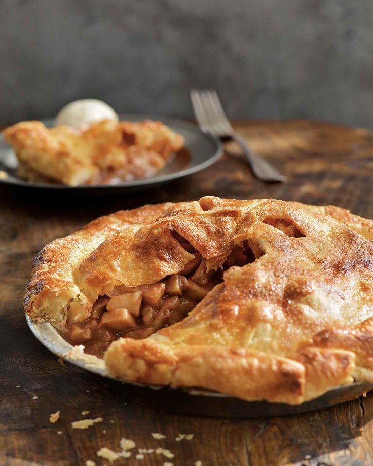 an apple pie on a wooden table next to a fork and plate with another pie in the background