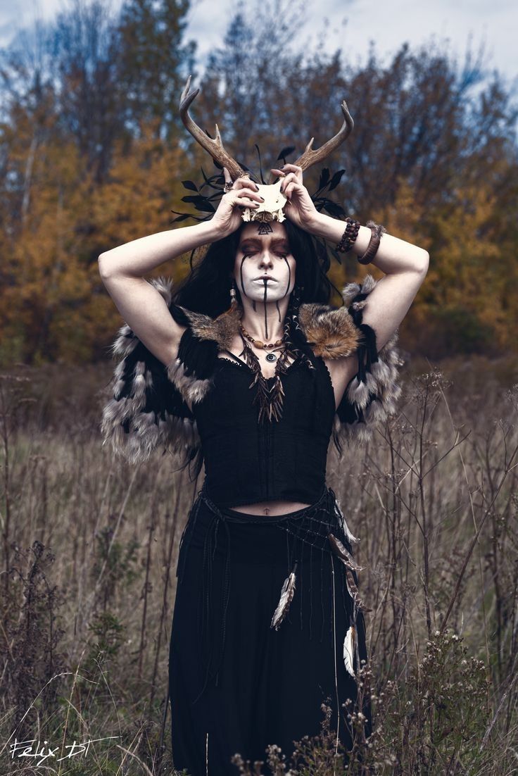 a woman with painted face and antlers on her head stands in tall grass, surrounded by trees